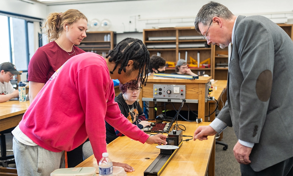 Students and faculty work on a project over a table in a classroom setting.
