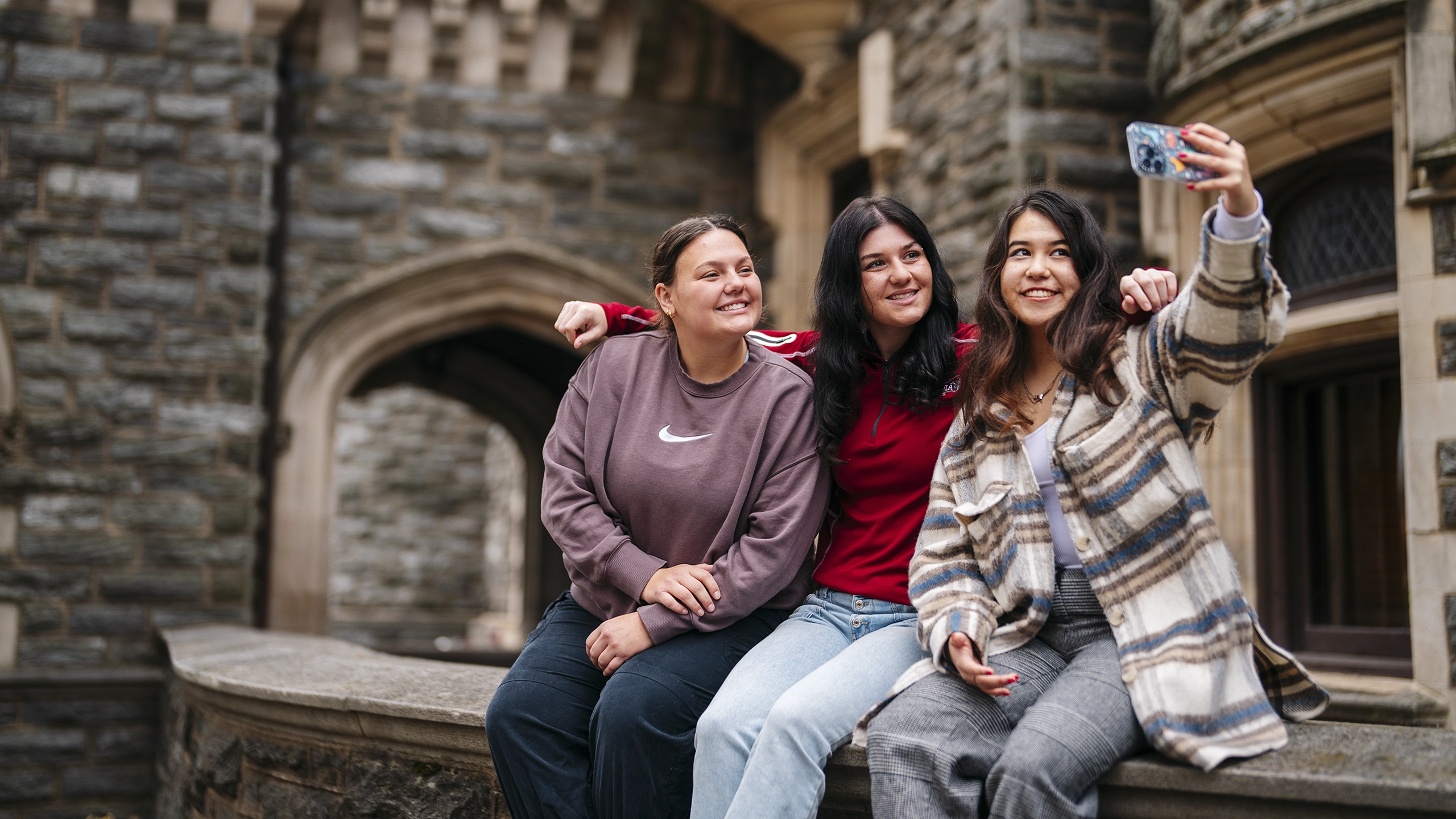 Three undergraduate students sit on a wall by Grey Towers Castle and take a selfie.