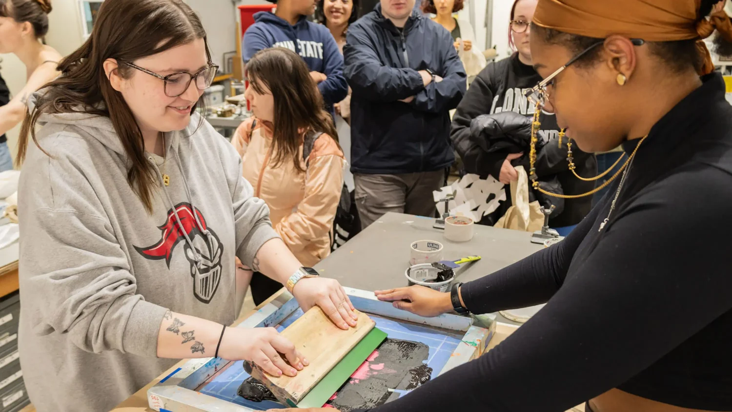 A student in a black shirt holds down a wooden frame while another student in an Arcadia sweatshirt presses ink into a print.