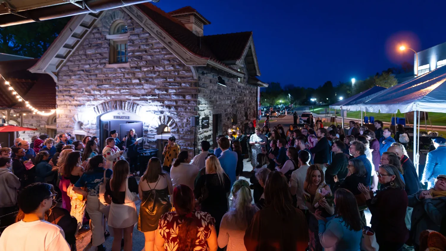 A crowd of students forms around a speaker with a microphone in front of the Theater at night on the Arcadia campus.