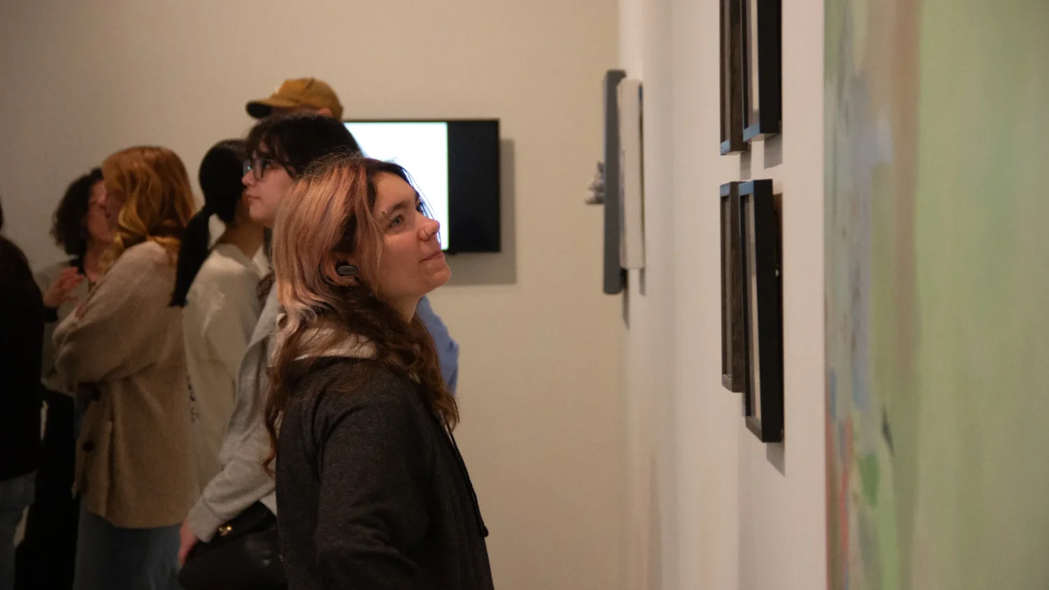 A student looks at artwork on a white wall with other students in the background.