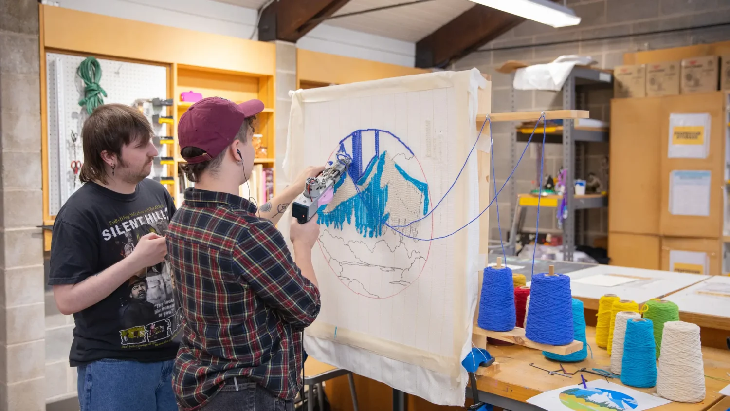 A student helps another student tuft blue yarn onto a canvas.