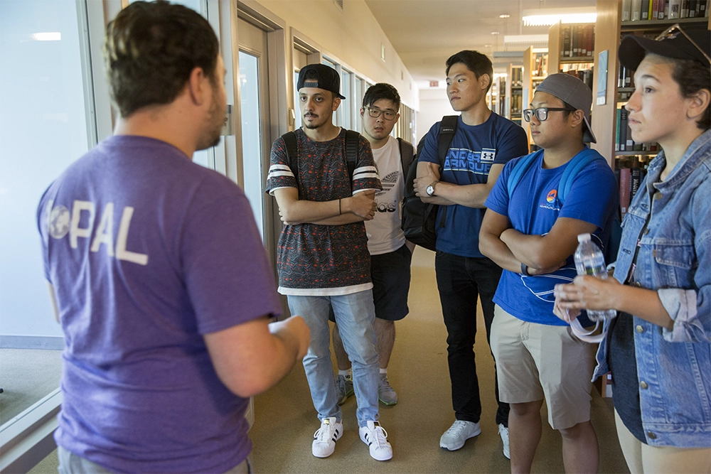 Four international students stand with their arms crossed in a library hallway and listen to a guide.