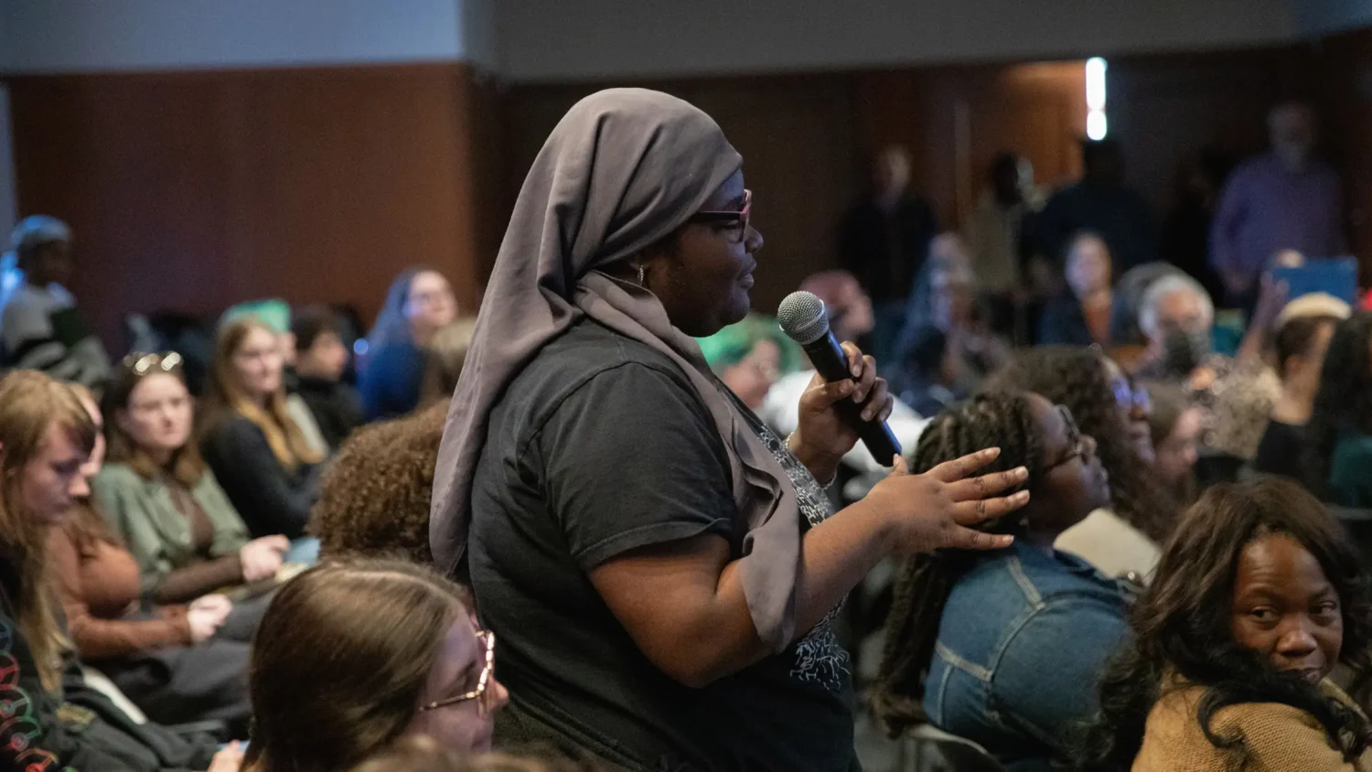 A student standing in the audience of a panel holds a microphone and gestures towards the panelists situated off-screen to the right.
