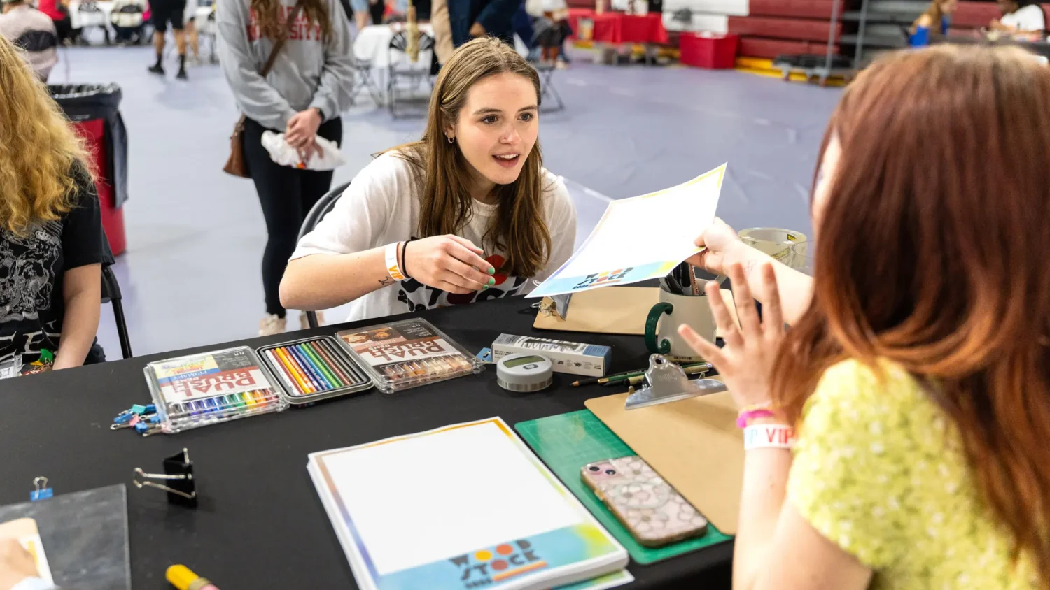 A student sitting at a table hands a piece of paper with a Woodstock footer to another student across from them. Various colored pencils and marker packages are laid out on the table.