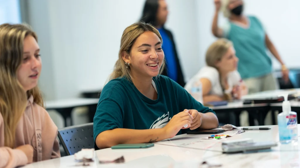 A student in a dark green shirt smiles while working at a table in a classroom.