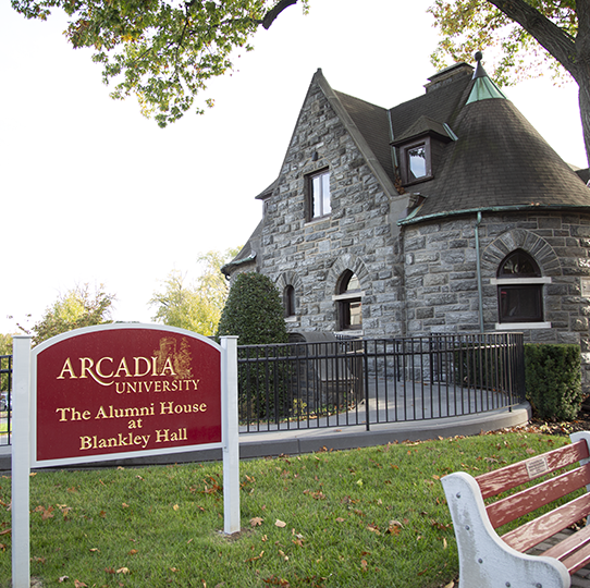 A red and gold lettered sign is on a green lawn outside of the Alumni House at Blankey Hall.