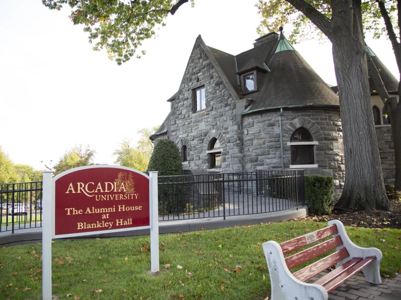A red and gold lettered sign is posted on a green lawn in front of the Alumni House at Blankey Hall.