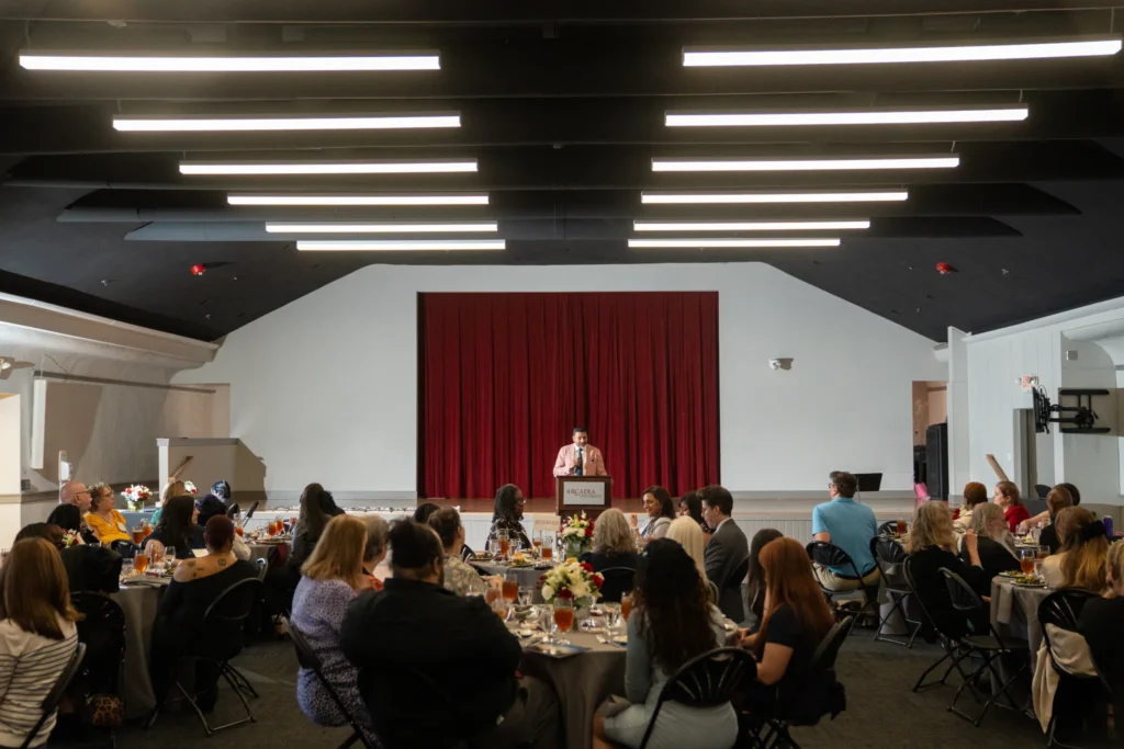 Man at a podium standing at the front of the auditorium addressing the attendees seated at their tables.