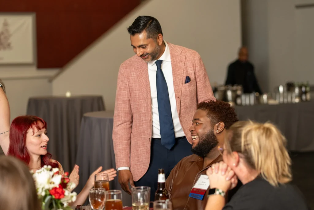 University President is smiling and laughing while standing around a tabled group of young alumni.