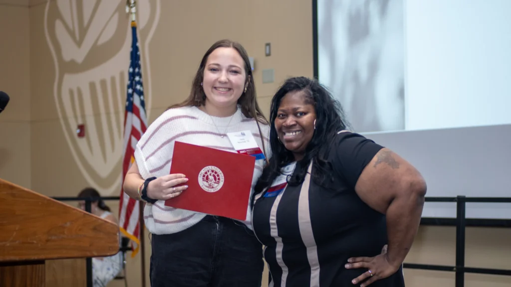 Two women on stage smiling. Woman on the left is holding her award.