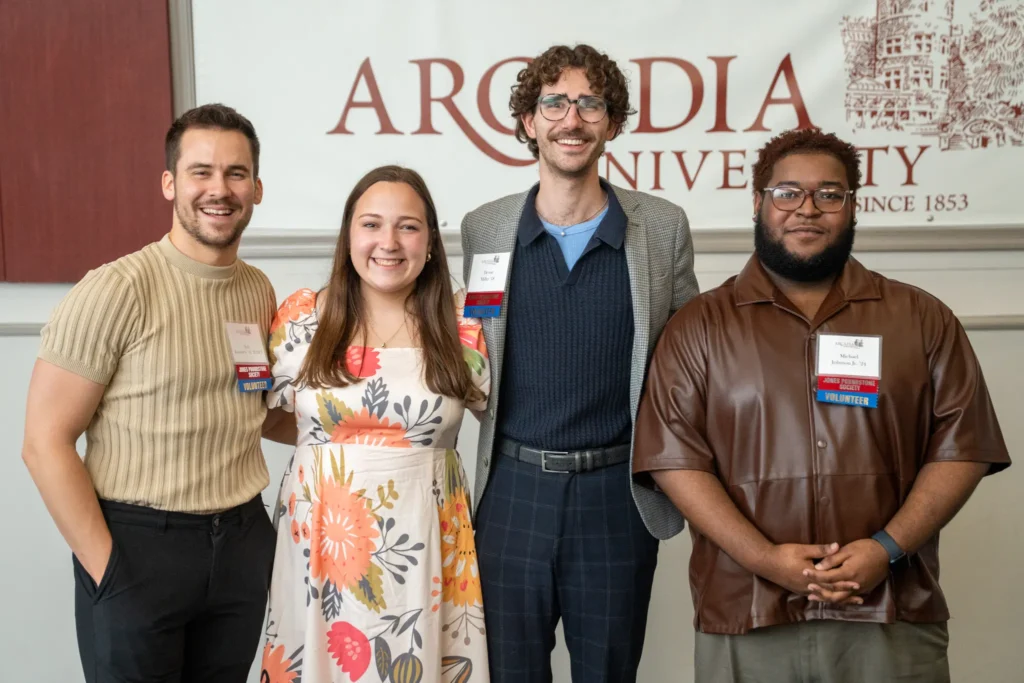 Four young alumni standing in front of an Arcadia University banner.