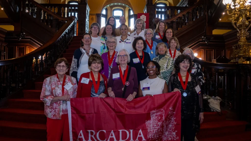 Group of alumni standing on the stairs of The Grey Towers Castle holding up an Arcadia University banner.