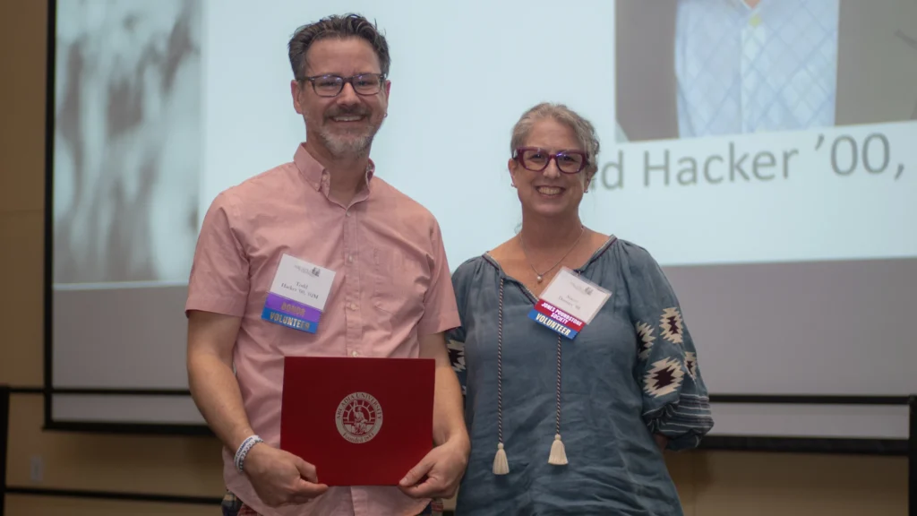 Man and woman stand on stage smiling after man won award. Man is holding award.