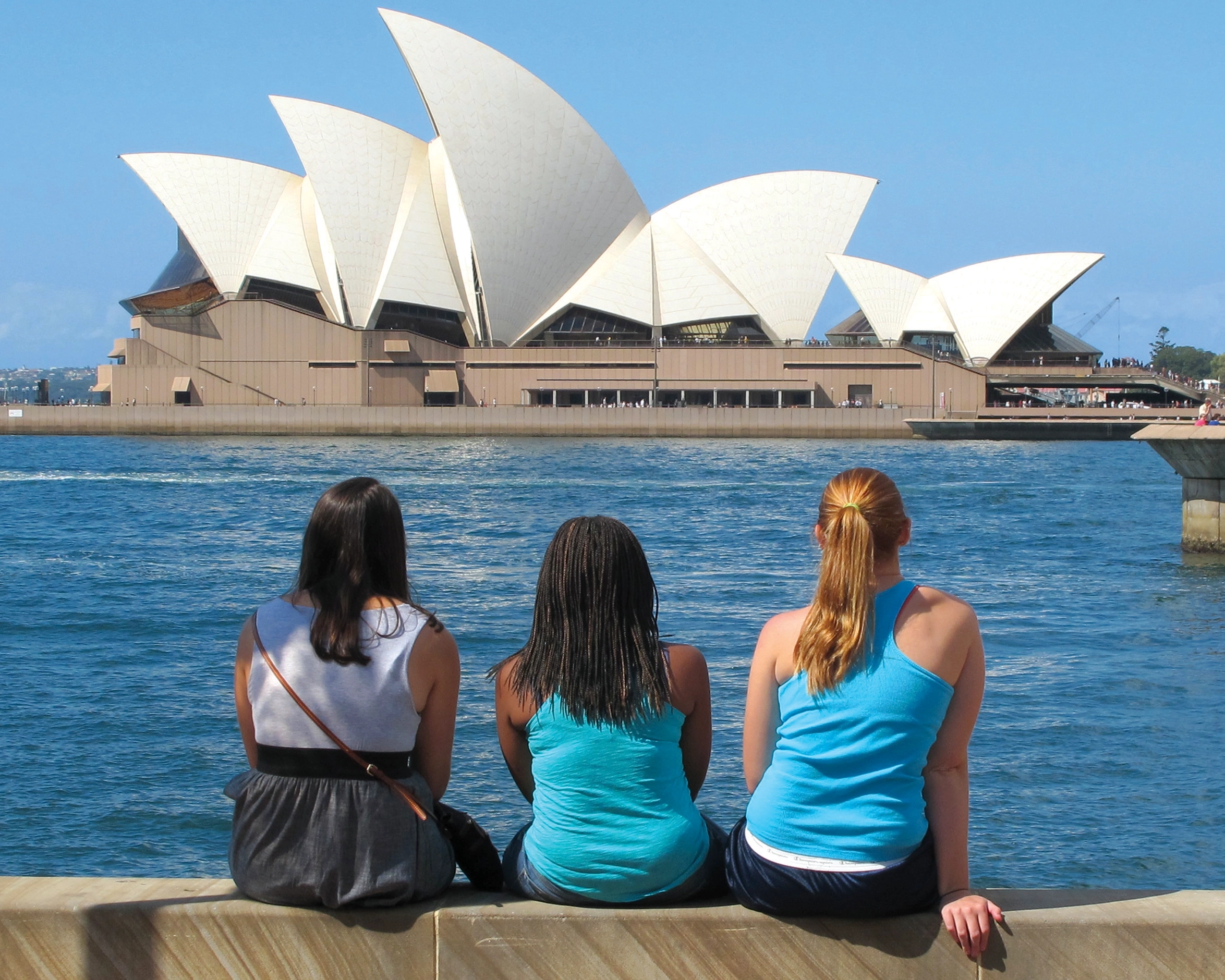 The Sydney Opera House is in the background as three students sit by the water.