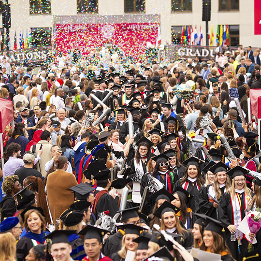 A crowd of students wear caps and gowns and celebrate as confetti drops during commencement.