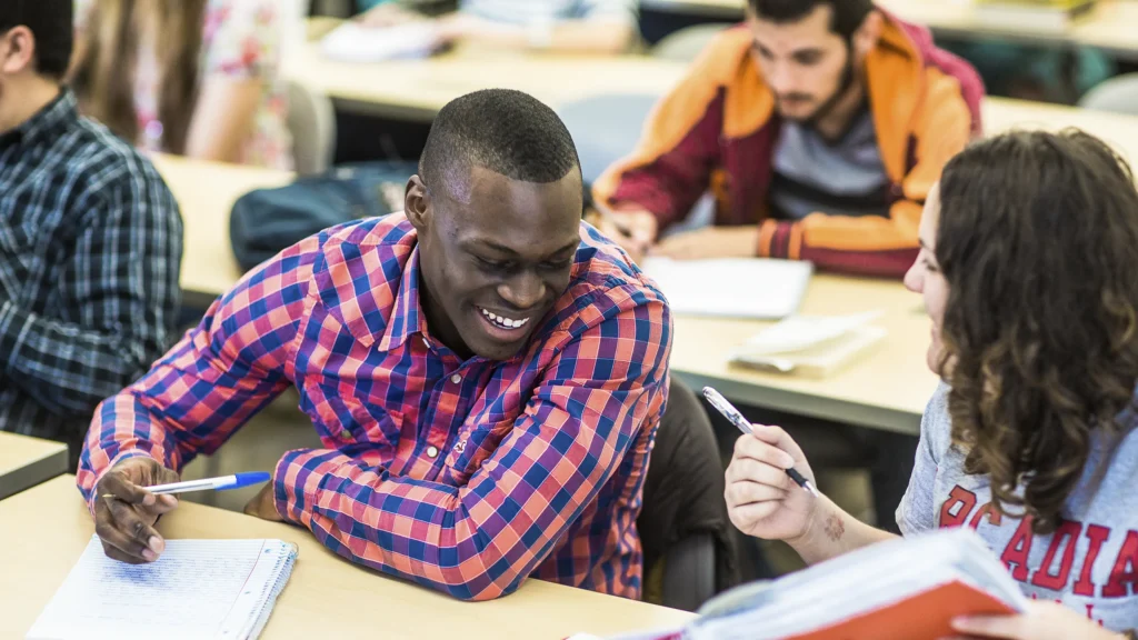 Undergraduate students sit in rows in a classroom as two take a break from their studies to chat.