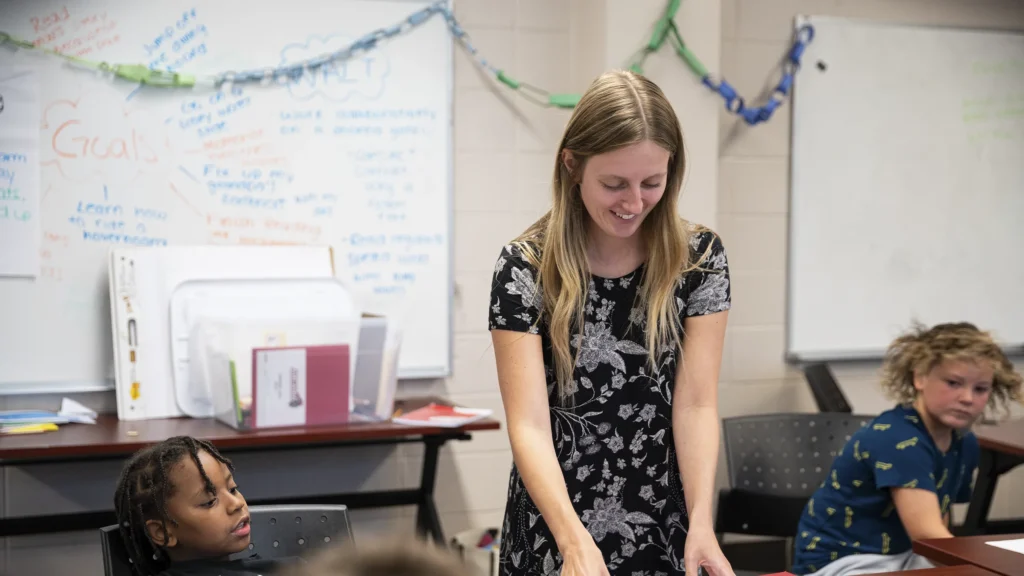 A teacher smiles as she helps elementary kids in a classrom.
