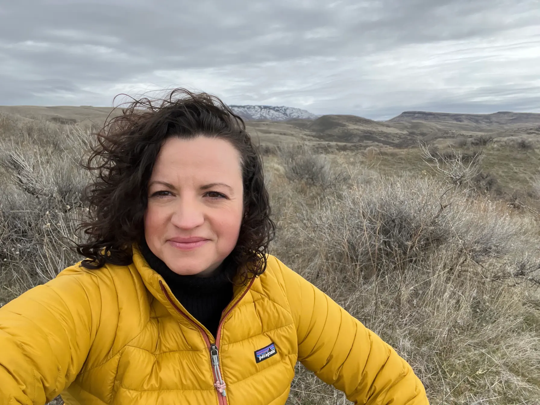 A woman in a yellow jacket takes a selfie at the top of a hill during a hiking trip.