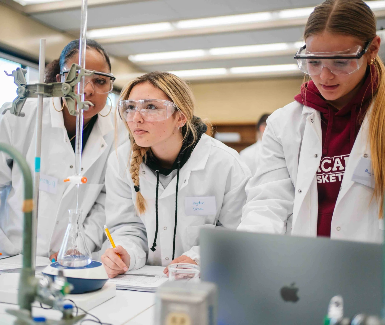 Three students in white lab coats and protective glasses perform an experiment in a lab.