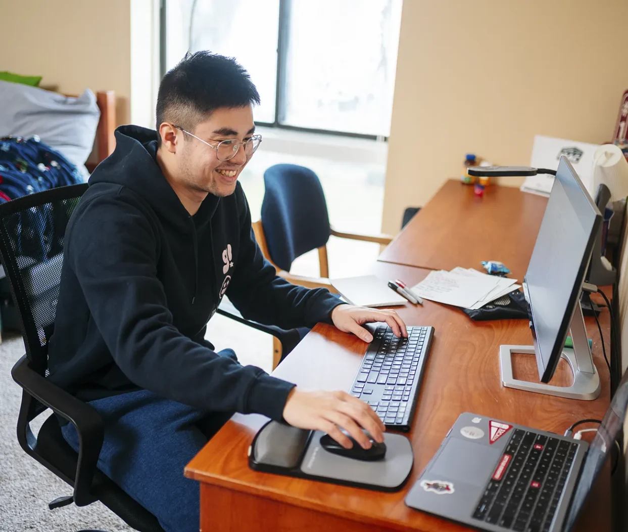 With a sunny window in the background, an undergraduate student works on a laptop in his dormroom.