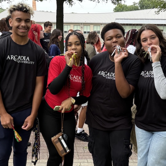 Four new Arcadia students (fall 2025) blow and hold noisemakers while looking at the camera