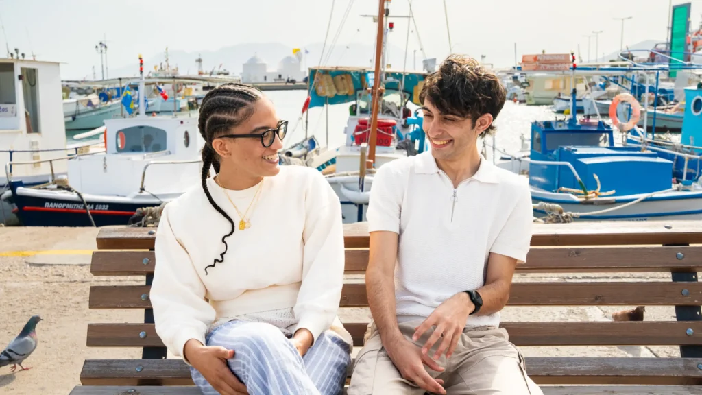 Two students sit on a bench and chat at a pier in Greece.