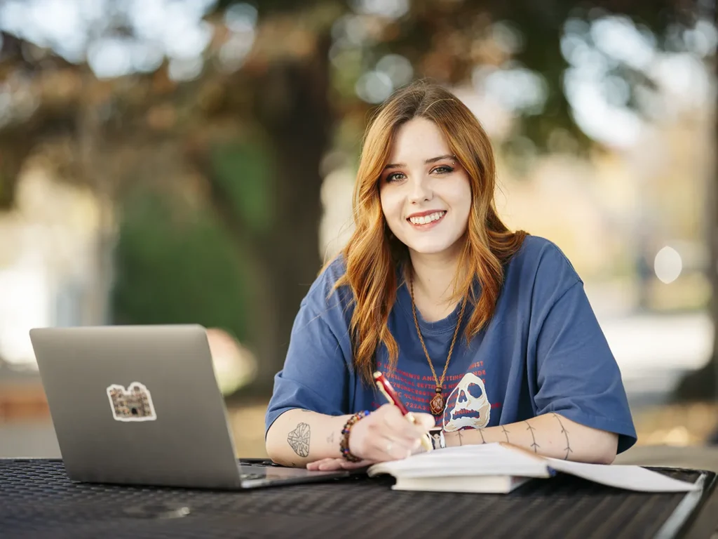 An undergraduate students smiles as she works on a laptop outside on campus.