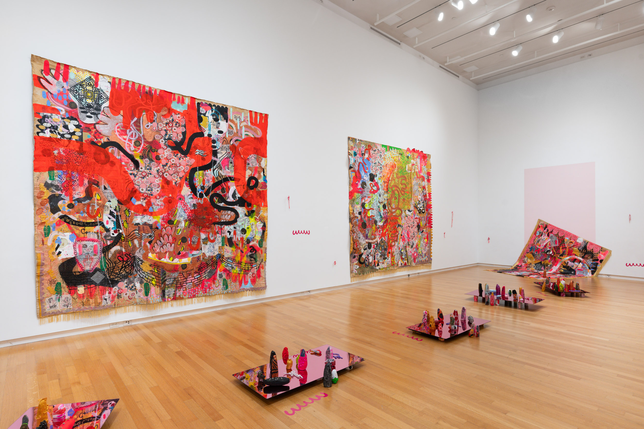 Installation view of Jackie Milad's work. Large gallery room with hardwood floors, small rectangular sculptures in the middle of the floor with small figurines placed on top. Two large multi media pieces on the walls, brightly colored with twisting black lines.