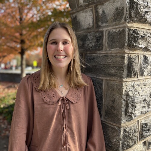 young woman standing next to a stone wall smiling on a fall day.