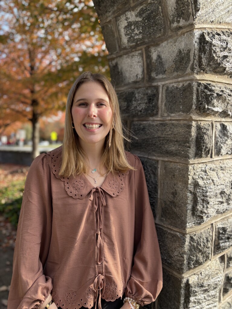 young woman standing next to a stone wall smiling on a fall day.