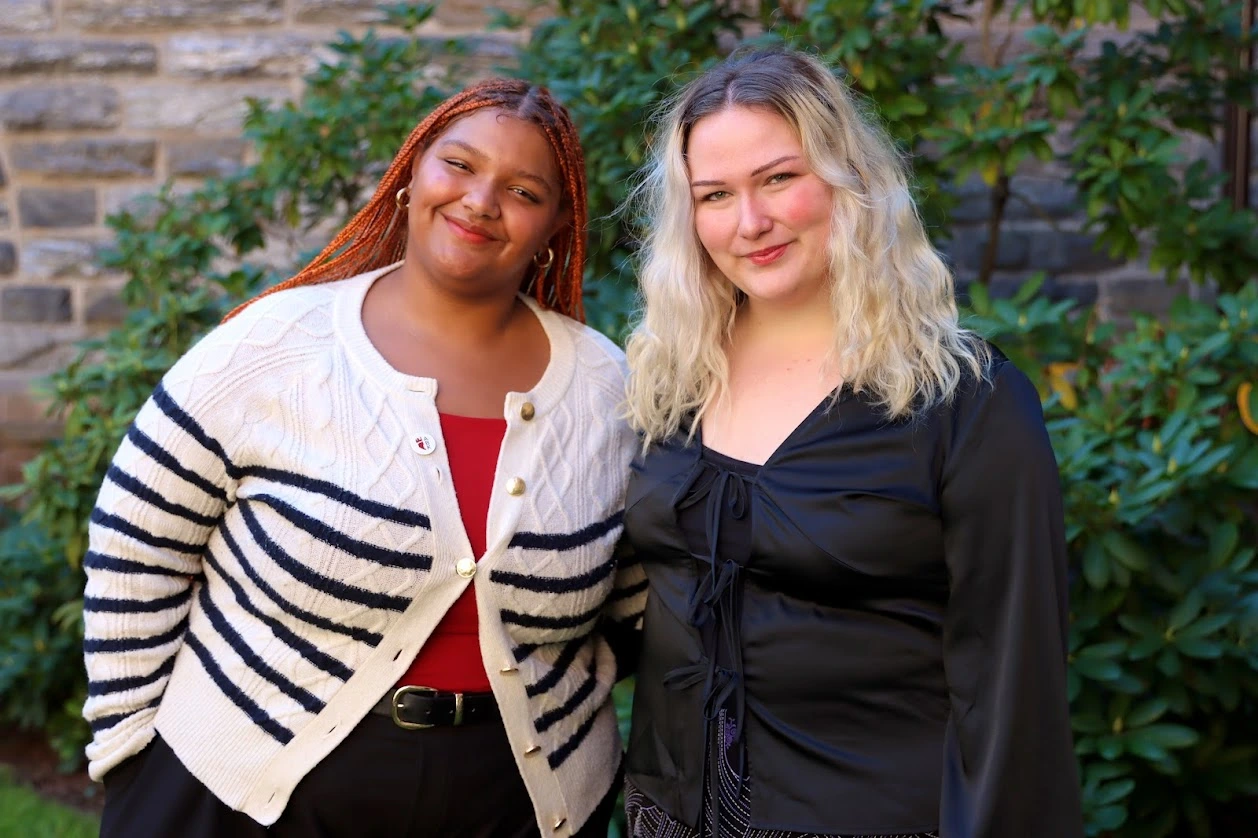 Two female students posed together and smiling with greenery as their backdrop.