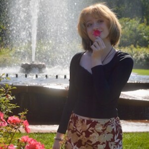 A student wearing a black top and red skirt hold a flower petal to her mouth