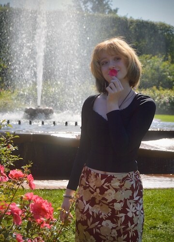 A student wearing a black top and red skirt hold a flower petal to her mouth