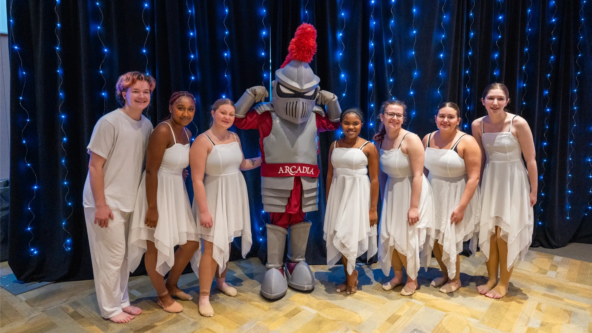 Students in white dresses pose for a photo with Archie the Knight.