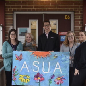 Five adults stand in front of the door to a building holding a blue illustrated poster that says "ASUA," which stands for A Step Up Academy.