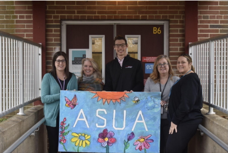 Five adults stand in front of the door to a building holding a blue illustrated poster that says "ASUA," which stands for A Step Up Academy.