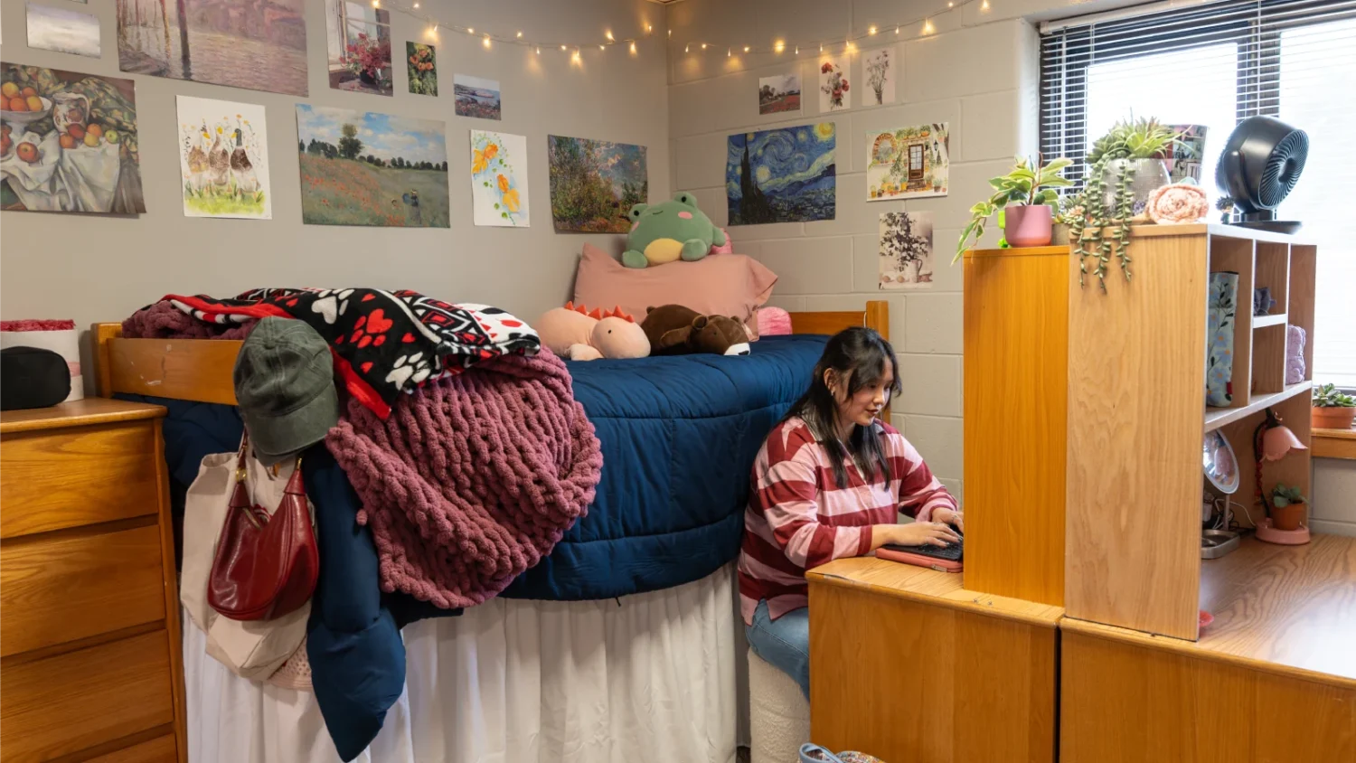 A student sits at their desk on their laptop while in their dorm room in the Knight Hall. A very elevated bed is positioned against the wall behind her.