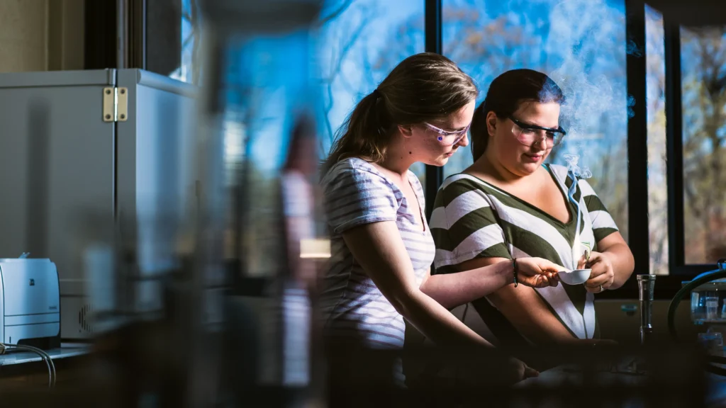 Two students wearing safety glasses work in a lab.