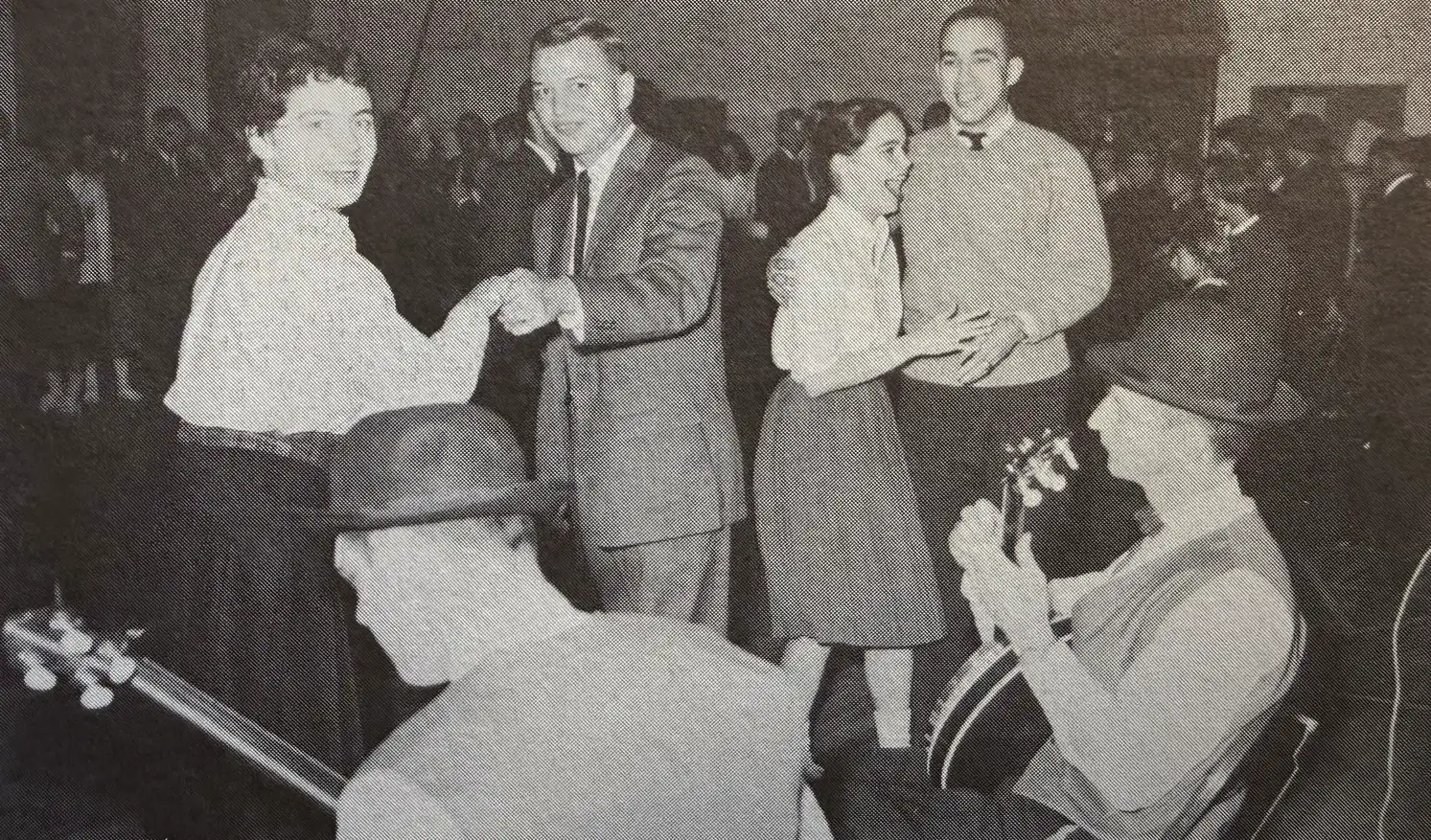 A black-and-white photo of students dancing at their school, with two men playing stringed instruments in the front.