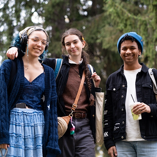 Three students wearing fall coats smile and pose in front of a green leafy tree.