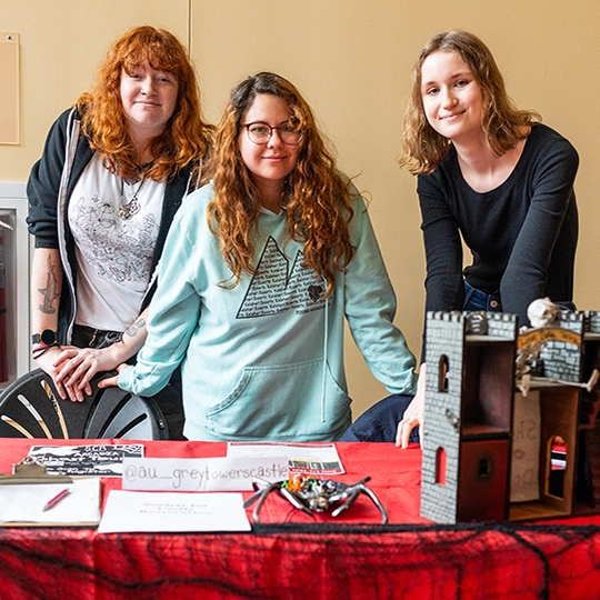 Three women stand behind a table covered with student information materials and university merchandise at an event.