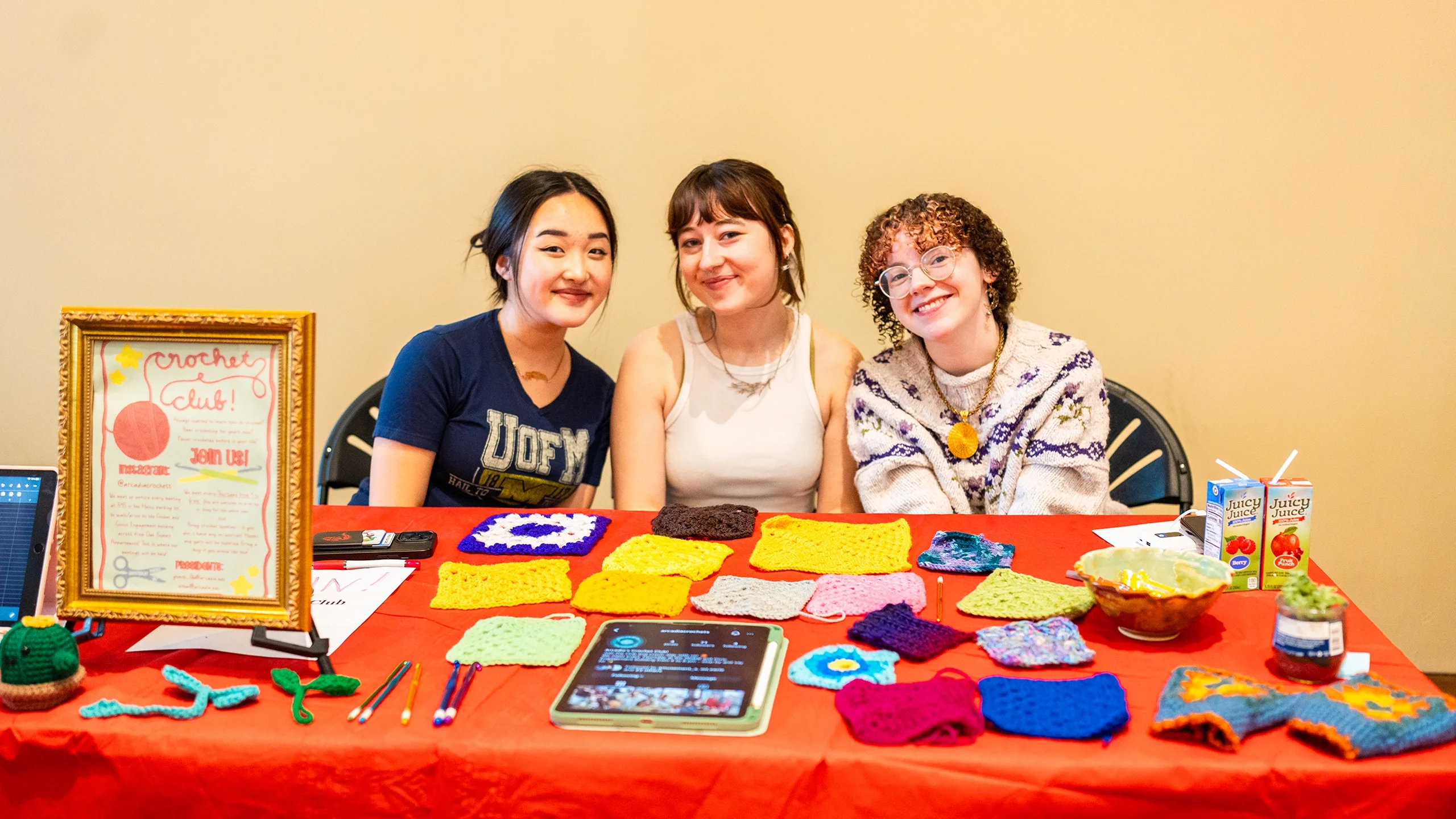 Three women sit behind a Crochet Club table at a campus activity fair, displaying their crochet work.