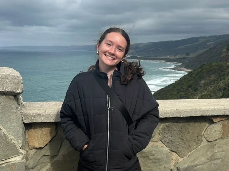 Callie Falzone '26 stands on a bridge and smiles while a beach is visible behind her.