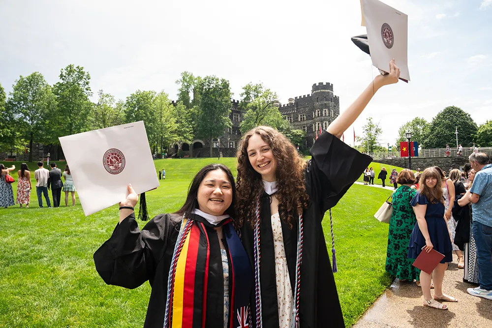 Two graduates wearing gowns hold their caps to celebrate together on graduation day.