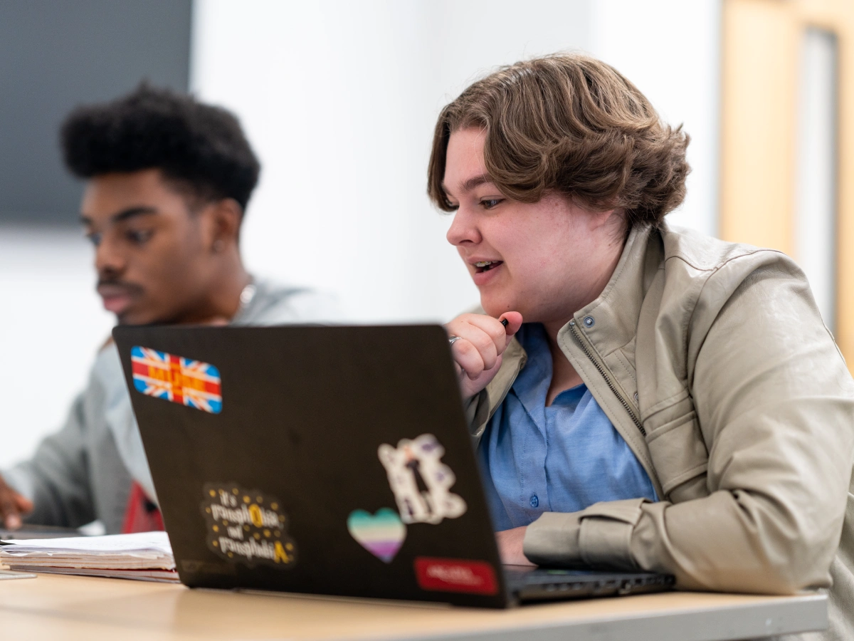 Two students sit at a desk facing to the left, while their laptops are open in front of them.