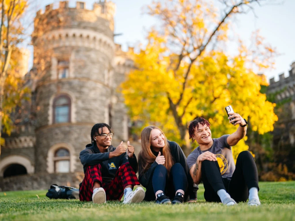 Three students sit in a field outside Grey Towers Castle posing for a selfie in the fall.