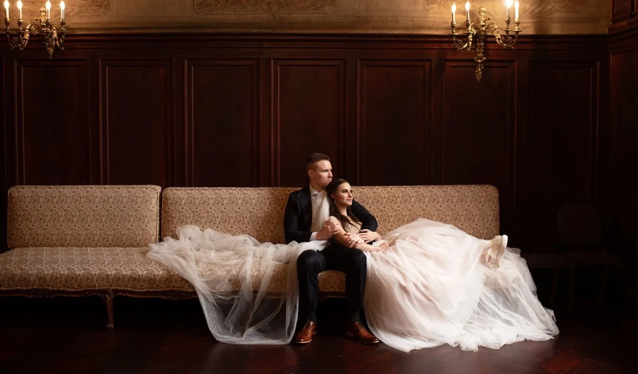 Couple in formal wedding attire reclining on a couch in a dimly lit room.