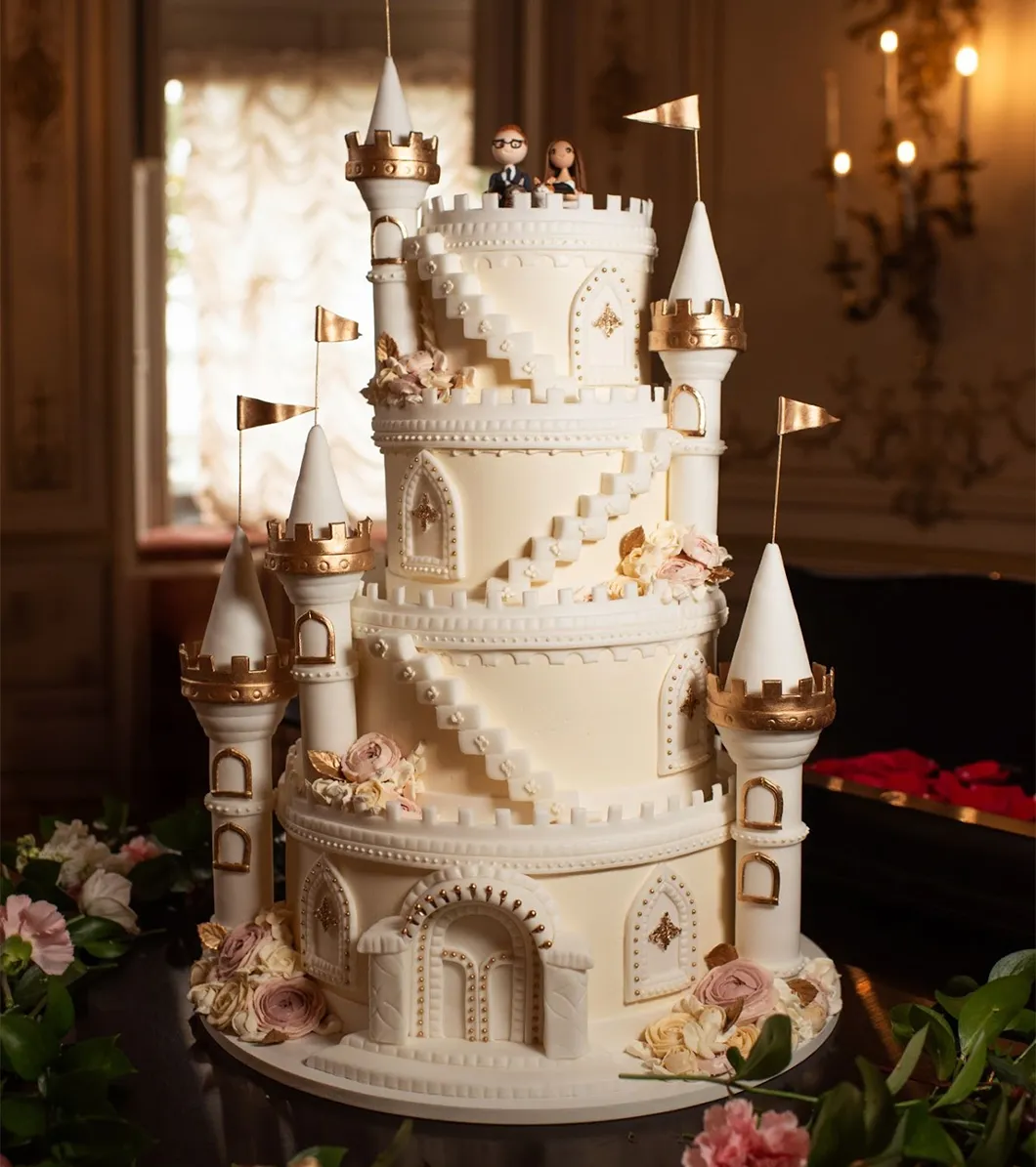 A four-tier wedding cake with castle turrets, small flags, gold trim, and a bride and groom topper.