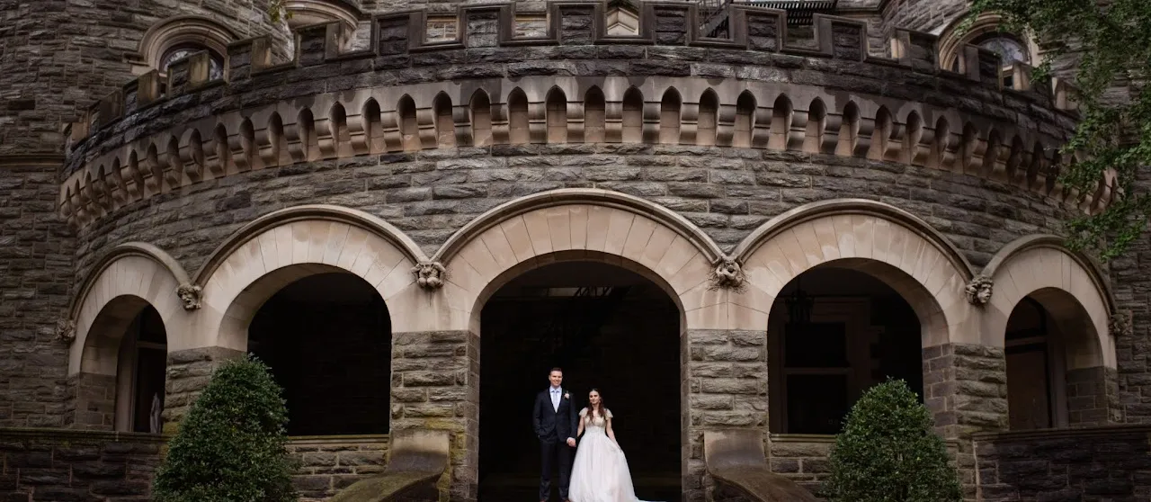 A formally dressed couple stands in front of the grey stone arched vestibule of the Grey Towers Castle.
